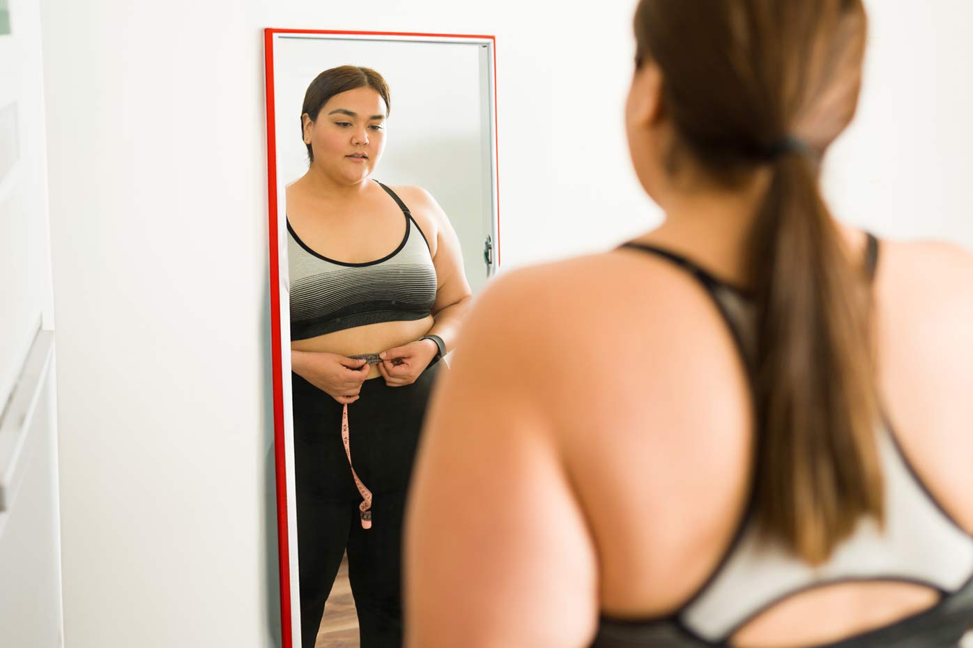 Larger woman measures her waist while looking at herself in the mirror