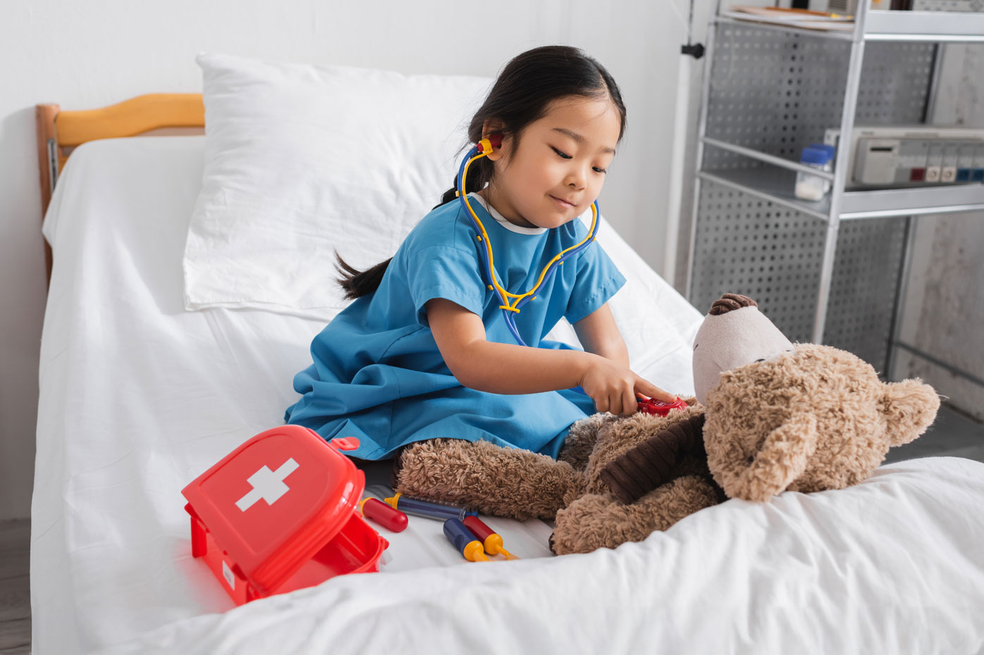 A little girl sits on a hospital bed playing with a doctor kit and a stuffed teddy bear