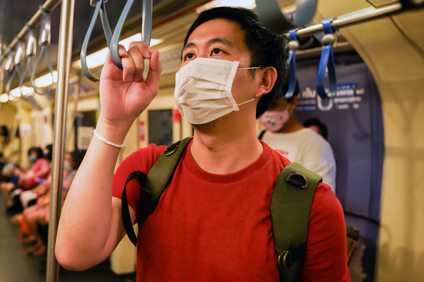 A man in a mask and red t-shirt holds a subway hand strap on a train filled with other masked passengers