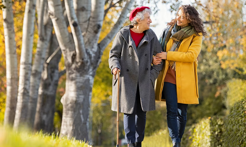 A senior woman in a gray coat and a red beret walks, smiling, arm in arm with a younger woman in a yellow coat