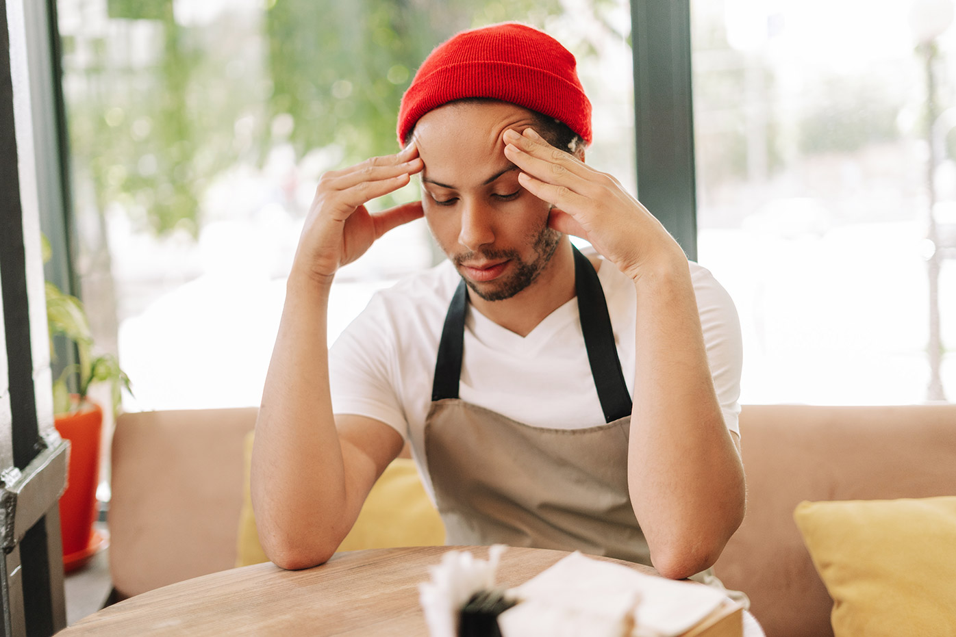 A seated man wearing red knit hat with his hands pressed to his forehead.