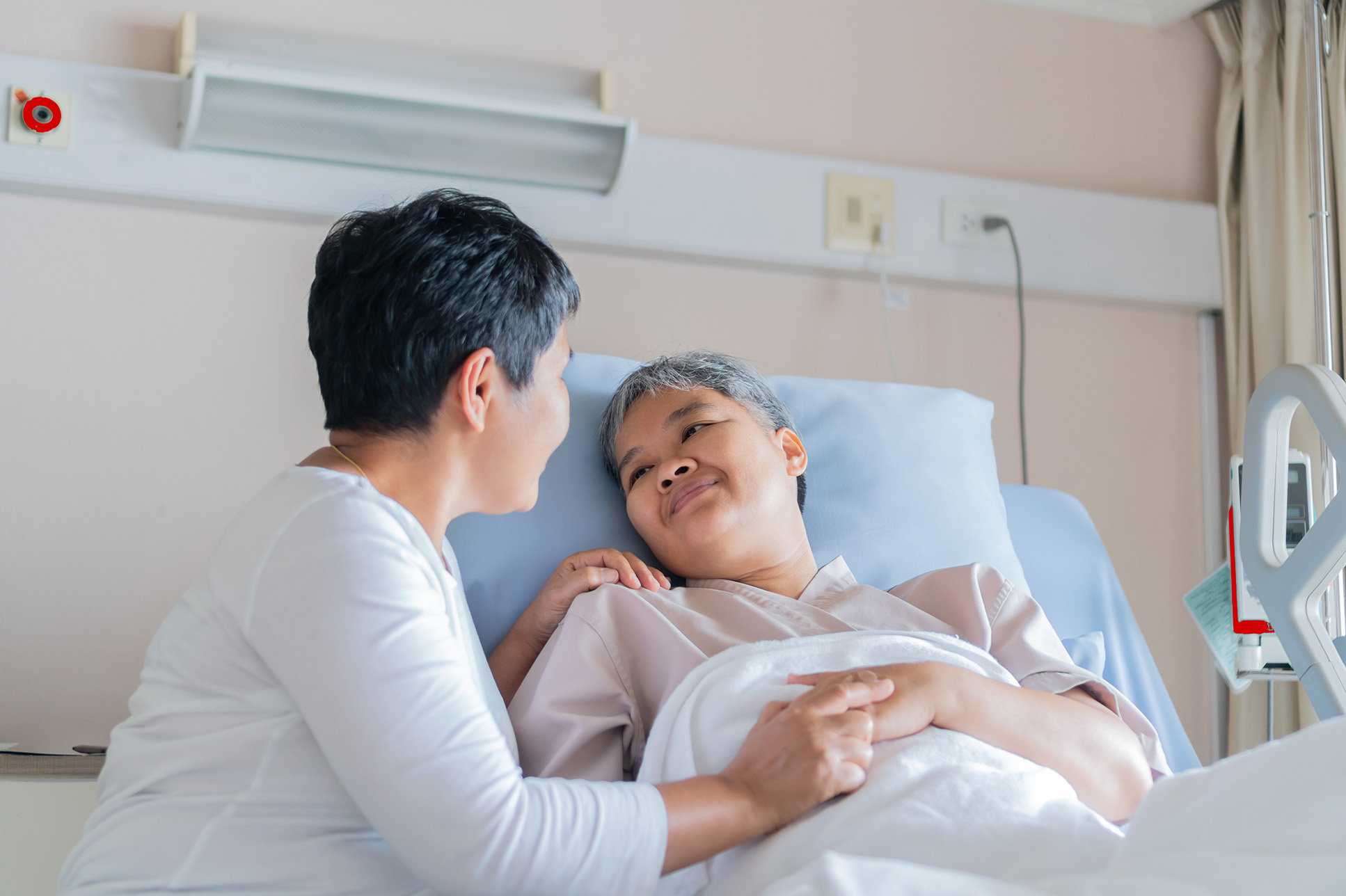 Asian woman holding mom's hand in hospital bed.